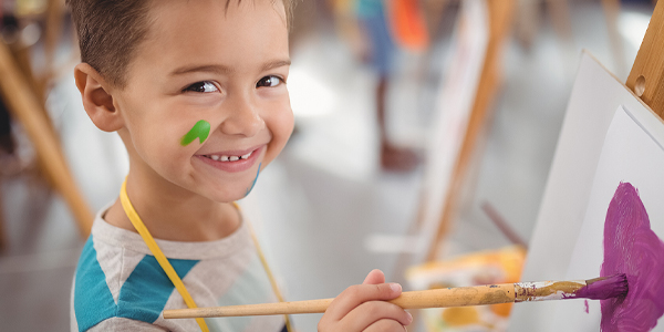 Aluno com o rosto pintado de tinta sorri ao fazer artes visuais que beneficiam a educação infantil no Colégio João Paulo I.