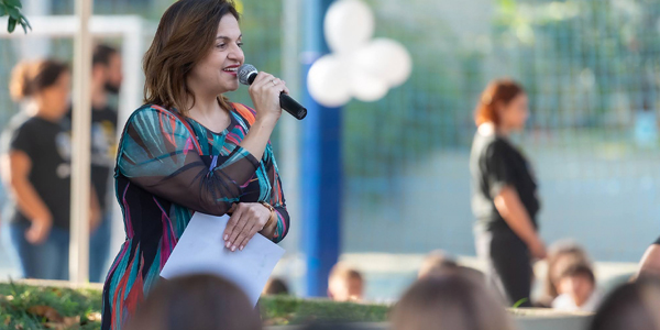 Educadora fala ao microfone em evento especial no Colégio João Paulo I