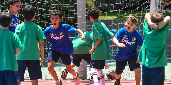 Grupo de alunos numa quadra participando de uma partida, um time com camisetas azuis e o outro com camisetas verdes. Muito empolgados com a Olijopa!